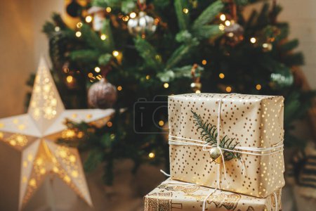 Foto de ¡Feliz Navidad! Elegantes regalos de Navidad en el árbol de Navidad con luces doradas. Regalos envueltos con papel dorado y rama de abeto bajo el árbol decorado en la habitación. Banner atmosférico, espacio de copia - Imagen libre de derechos