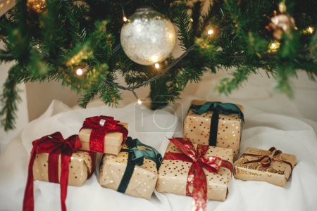 Téléchargez les photos : Cadeaux de Noël élégants sous l'arbre de Noël avec des lumières dorées. Cadeaux de Noël enveloppés dans du papier doré avec des rubans rouges et verts dans une salle moderne festive. Joyeux Noël et Joyeuses fêtes - en image libre de droit