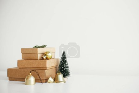 Téléchargez les photos : Élégantes boîtes-cadeaux éco-Noël sur table blanche. Cadeaux de Noël artisanaux simples avec des boules d'or, arbre et branche de sapin sur bois rustique. Joyeux Noël ! Vacances écologiques, espace pour le texte - en image libre de droit
