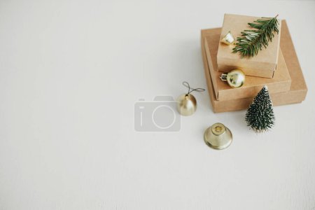 Foto de Elegantes cajas de regalo eco Navidad en mesa blanca. Simple navidad artesanal presenta con bolas de oro, árbol y rama de abeto en madera rústica. ¡Feliz Navidad! Vacaciones ecológicas, espacio para el texto - Imagen libre de derechos