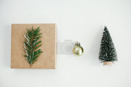 Photo for Merry Christmas! Stylish eco christmas gift box, golden bauble and tree decoration on white table flat lay. Simple craft christmas present on rustic wood. Zero waste holiday. Minimal still life - Royalty Free Image