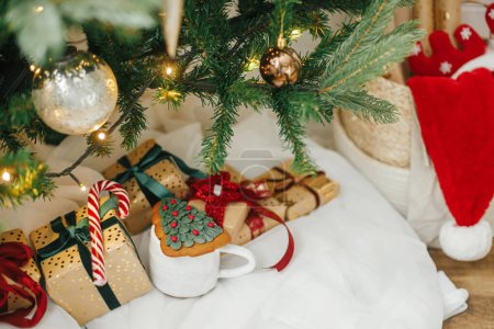Foto de ¡Feliz Navidad! Elegantes regalos de Navidad bajo el árbol de Navidad con luces doradas. Regalos de Navidad envueltos en papel dorado con cintas rojas y verdes, bastón de caramelo, galleta en habitación festiva - Imagen libre de derechos
