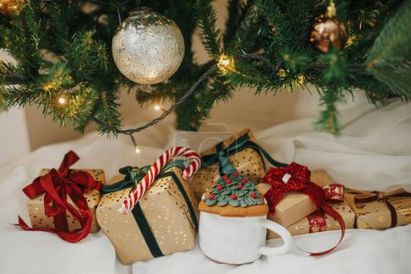 Foto de Elegantes regalos de Navidad bajo el árbol de Navidad con luces doradas. Envuelto Navidad presenta en papel dorado con cintas rojas y verdes, bastón de caramelo, galleta en habitación festiva. Feliz Navidad. - Imagen libre de derechos