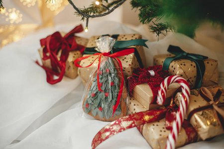 Foto de ¡Feliz Navidad! Elegantes regalos de Navidad con galleta y bastón de caramelo bajo el árbol de Navidad con luces doradas. Regalos de Navidad envueltos en papel dorado con cintas rojas y verdes en sala festiva - Imagen libre de derechos