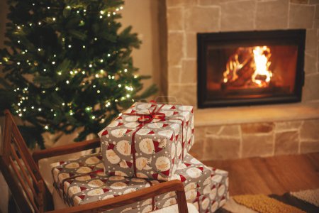 Foto de ¡Feliz Navidad! Elegantes regalos de Navidad envueltos en el fondo del árbol con luces y chimenea en habitación rústica por la noche. Acogedoras vacaciones de invierno, víspera de Navidad. Copiar espacio - Imagen libre de derechos