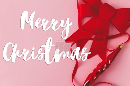 Foto de Feliz Navidad texto en el arco rojo de Navidad, bauble y cinta sobre fondo rosa plana laico. Tarjeta de felicitación de la temporada. Signo manuscrito - Imagen libre de derechos