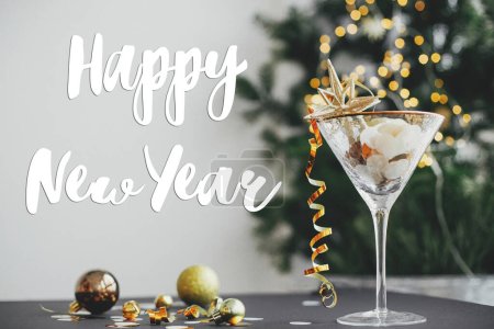 Foto de Feliz año nuevo signo de texto en la elegante estrella de brillo de Navidad en copa de champán, confeti de oro y adornos en el fondo de luces festivas. Tarjeta de felicitación de temporada - Imagen libre de derechos