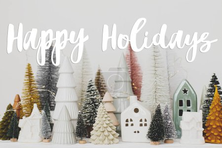 Photo for Happy Holidays text sign on stylish little Christmas trees and houses decorations on white table. Modern christmas scene, miniature cozy snowy village. Season's greeting card - Royalty Free Image