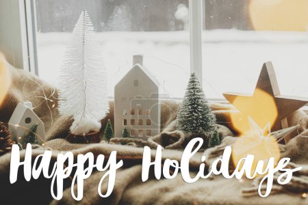 Foto de Cartel de texto de Happy Holidays en un elegante árbol de Navidad, luces, casita y estrella de madera en una acogedora manta en la ventana. Tarjeta de felicitación de la temporada. Invierno hygge casa - Imagen libre de derechos