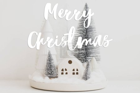 Photo for Merry Christmas text on stylish little Christmas trees and house on white table. Season's greeting card. Cozy christmas magical scene, miniature snowy village. Handwritten sign - Royalty Free Image