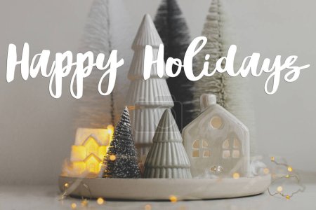 Photo for Happy Holidays text on stylish little Christmas trees and house on white table. Season's greeting card. Cozy christmas magical scene, miniature snowy village with lights. Handwritten sign - Royalty Free Image