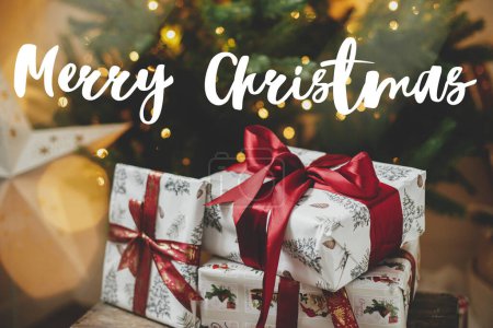 Téléchargez les photos : Joyeux Noël signe de texte sur le cadeau de Noël élégant sur fond d'arbre de Noël avec des lumières dorées dans la salle du soir. La carte de voeux de la saison. Souhaits atmosphériques - en image libre de droit