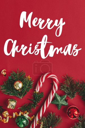 Photo for Merry Christmas text on christmas baubles, fir branches, confetti and candy cane on red background flat lay. Season's greeting card. Handwritten sign - Royalty Free Image