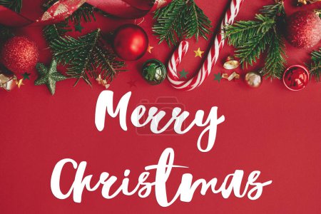 Photo for Merry Christmas text on christmas baubles, fir branches, confetti and candy cane on red background flat lay. Season's greeting card. Handwritten sign - Royalty Free Image