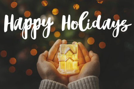 Photo for Happy Holidays text sign on christmas glowing house in hands on background of illuminated christmas lights bokeh. Season's greeting card. Christmas postcard - Royalty Free Image