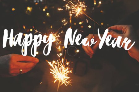 Photo for Happy New Year text sign on hands holding fireworks against christmas lights in dark room. Season's greeting card. Friends celebrating with burning sparklers - Royalty Free Image