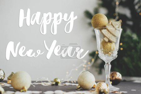 Foto de Feliz año nuevo cartel de texto en bolas de brillo dorado en copa de champán y decoraciones de Navidad brillantes en la mesa oscura. Tarjeta de felicitación de temporada - Imagen libre de derechos