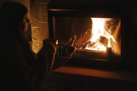 Photo for Woman holding cup of tea and warming up hands at cozy fireplace in dark evening room. Fireplace heating in house, electricity blackout. Atmospheric time at fireside in home - Royalty Free Image