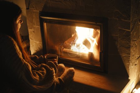 Photo for Stylish woman warming up hands and feet at cozy fireplace with tea in evening. Fireplace heating in house, electricity blackout. Atmospheric time at fireside in home - Royalty Free Image