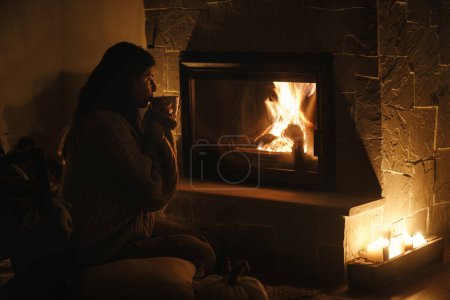 Téléchargez les photos : Femme buvant une tasse de thé chaud, assis à la cheminée confortable dans la salle du soir sombre. Chauffage par cheminée dans la maison, panne d'électricité. Temps atmosphérique au coin du feu à la maison - en image libre de droit