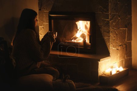 Photo for Stylish woman with warm cup of tea sitting at cozy fireplace in dark evening room. Fireplace heating in house, electricity blackout. Atmospheric time at fireside in home - Royalty Free Image