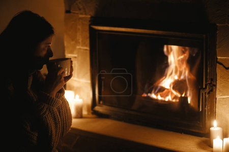 Photo for Woman drinking warm cup of tea, sitting at cozy fireplace in dark evening room. Fireplace heating in house, electricity blackout. Atmospheric time at fireside in home - Royalty Free Image
