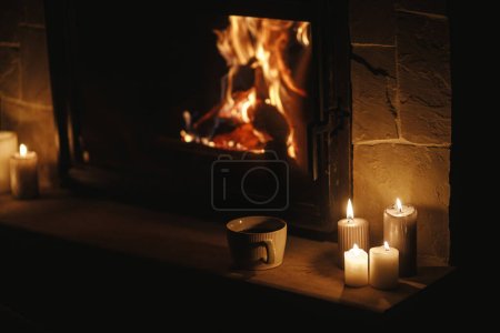 Téléchargez les photos : Tasse chaude de thé et de bougies à la cheminée à combustion confortable dans la salle sombre du soir. Chauffage par cheminée dans la maison, panne d'électricité. Temps atmosphérique au coin du feu à la maison - en image libre de droit