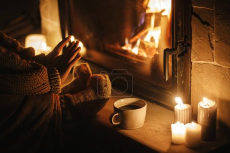 Foto de Mujer con estilo que calienta las manos y los pies en la acogedora chimenea con té por la noche. Chimenea calefacción en casa, apagón eléctrico. Tiempo atmosférico en la chimenea en casa - Imagen libre de derechos