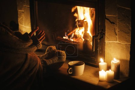 Stilvolle Frau wärmt Hände und Füße am gemütlichen Kamin mit Tee am Abend. Kaminheizung im Haus, Stromausfall. Stimmungsvolle Zeit am heimischen Kamin