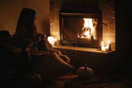 Photo for Stylish woman with warm cup of tea sitting at cozy fireplace in dark evening room. Fireplace heating in house, electricity blackout. Atmospheric time at fireside in home - Royalty Free Image