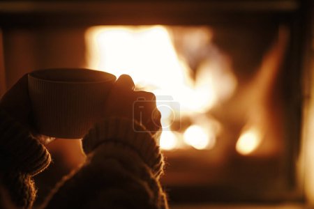 Photo for Woman holding cup of tea and warming up hands at cozy fireplace in dark evening room, close up. Fireplace heating in house, electricity blackout. Atmospheric time at fireside in home - Royalty Free Image