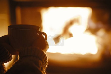 Foto de Mujer sosteniendo la taza de té y calentando las manos en la acogedora chimenea en la sala de noche oscura, de cerca. Chimenea calefacción en casa, apagón eléctrico. Tiempo atmosférico en la chimenea en casa - Imagen libre de derechos