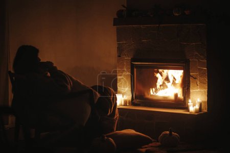Téléchargez les photos : Silhouette de femme avec une tasse de thé chaud assis à la cheminée confortable dans la salle du soir sombre. Chauffage par cheminée dans la maison, panne d'électricité. Temps atmosphérique au coin du feu à la maison - en image libre de droit