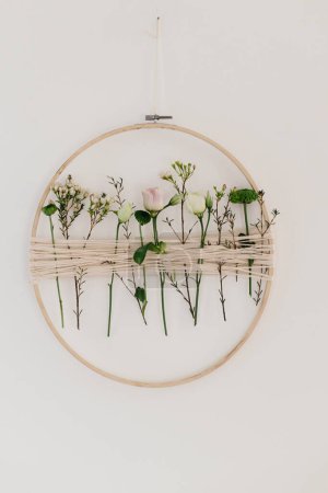 Photo for Stylish spring wreath with beautiful fresh flowers. Wooden hoop with flowers and thread hanging on white wall. Modern and creative floral handmade decor - Royalty Free Image