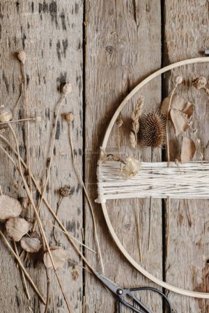 Téléchargez les photos : Making stylish boho autumn wreath with dry flowers flat lay. Wooden hoop, thread, scissors, and dry herbs on rustic wooden background. Modern floral arrangement and handmade decor - en image libre de droit
