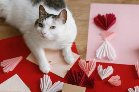 Foto de Cute cat lying at red and pink hearts, gift box, envelope, scissors and paper on wooden table. Valentine holiday preparations. Happy Valentine's day! Adorable kitty helper. Pet and love - Imagen libre de derechos
