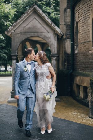 Photo for Beautiful emotional wedding couple kissing and walking in european city. Provence wedding. Stylish happy bride and groom walking on background of church in sunny street - Royalty Free Image