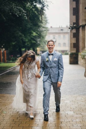 Foto de Elegante novia feliz y novio caminando sobre el fondo de la antigua iglesia en la calle lluviosa. Boda de Provenza. Hermosa pareja de boda emocional sonriendo bajo la lluvia en la ciudad europea. - Imagen libre de derechos