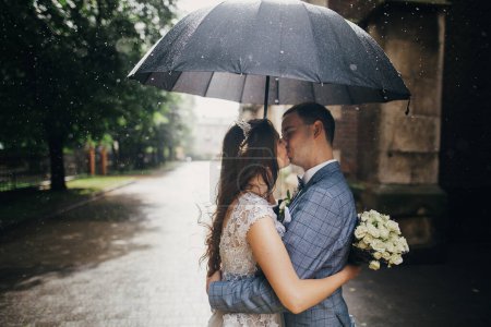 Photo for Stylish bride with bouquet and groom kissing under umbrella on background of old church in rain. Provence wedding. Beautiful wedding couple embracing under black umbrella in rainy street. Romantic moment - Royalty Free Image