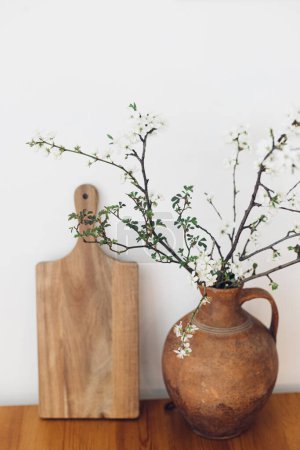 Téléchargez les photos : Blooming cherry branches in old vase and wooden board on table against white wall. Spring flowers in kitchen still life. Simple countryside living, home rustic decor. Hello spring - en image libre de droit