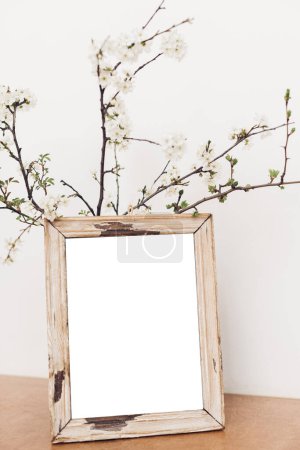 Photo for Photo frame mockup and blooming cherry branches on wooden table against white wall. Empty picture frame template and spring flowers. Simple countryside living. Space for text - Royalty Free Image