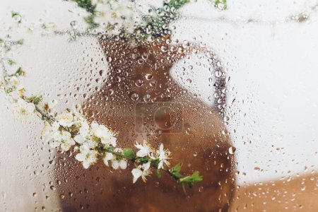 Foto de Cherry blooming branch in vase behind glass with water drops. Creative abstract image of spring flowers. Simple aesthetic wallpaper, wet rainy flowers. Floral rustic still life, simple living - Imagen libre de derechos