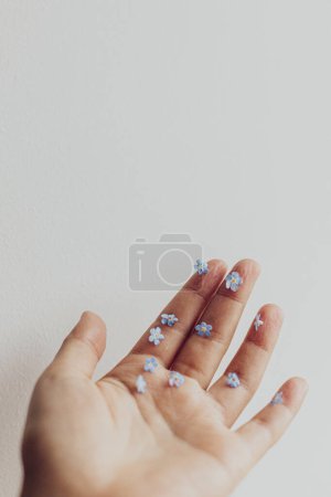 Foto de Hello spring. Simple aesthetic image. Cute little blue flowers on hand and fingers against white wall. Delicate forget me not petals on palm.  Skin care and organic cosmetics concept - Imagen libre de derechos
