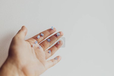 Photo for Cute little blue flowers on hand and fingers against white wall. Skin care and organic cosmetics concept. Delicate forget me not petals on palm. Hello spring. Simple aesthetic image - Royalty Free Image