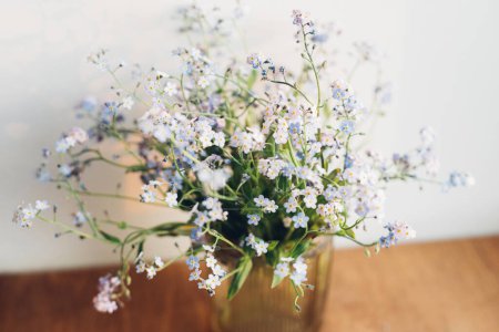 Photo for Beautiful little blue flowers in vase in warm evening sunlight on rustic wooden table. Delicate myosotis petals, forget me not spring flowers. Simple countryside living, home decor - Royalty Free Image