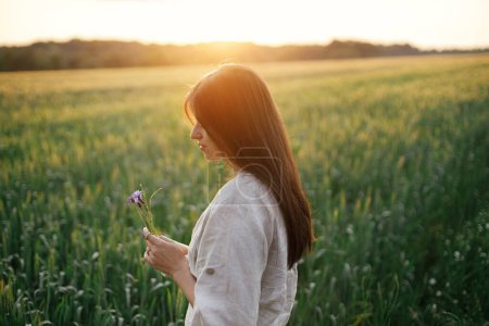 Téléchargez les photos : Woman relaxing in wheat field in warm sunset light. Tranquil atmospheric moment. Portrait of young female in rustic dress holding wildflowers in hands in evening summer countryside - en image libre de droit