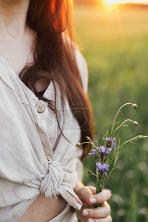 Téléchargez les photos : Woman holding wildflowers in wheat field in warm sunset light. Close up of young female in rustic dress with flowers in hands in evening summer countryside. Tranquil atmospheric moment - en image libre de droit