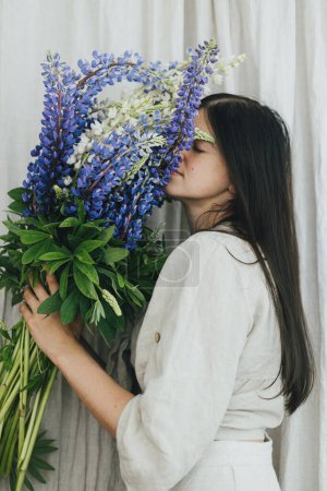 Téléchargez les photos : Stylish woman holding lupine bouquet in rustic room. Gathering and arranging summer wildflowers at home in countryside. Portrait of young female in linen dress posing with lupine flowers - en image libre de droit