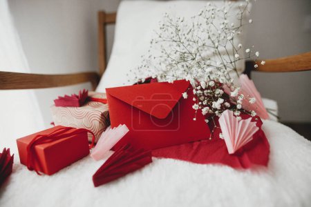 Foto de Happy valentines day! Modern gifts, red envelope, hearts and white flowers on cozy armchair. Valentine morning surprise. Valentines day gift - Imagen libre de derechos