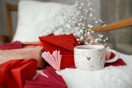 Foto de Cute stylish cup of tea with heart on cozy modern armchair with gifts, red envelope and white flowers. Valentine morning surprise for beloved. Happy valentines day! - Imagen libre de derechos